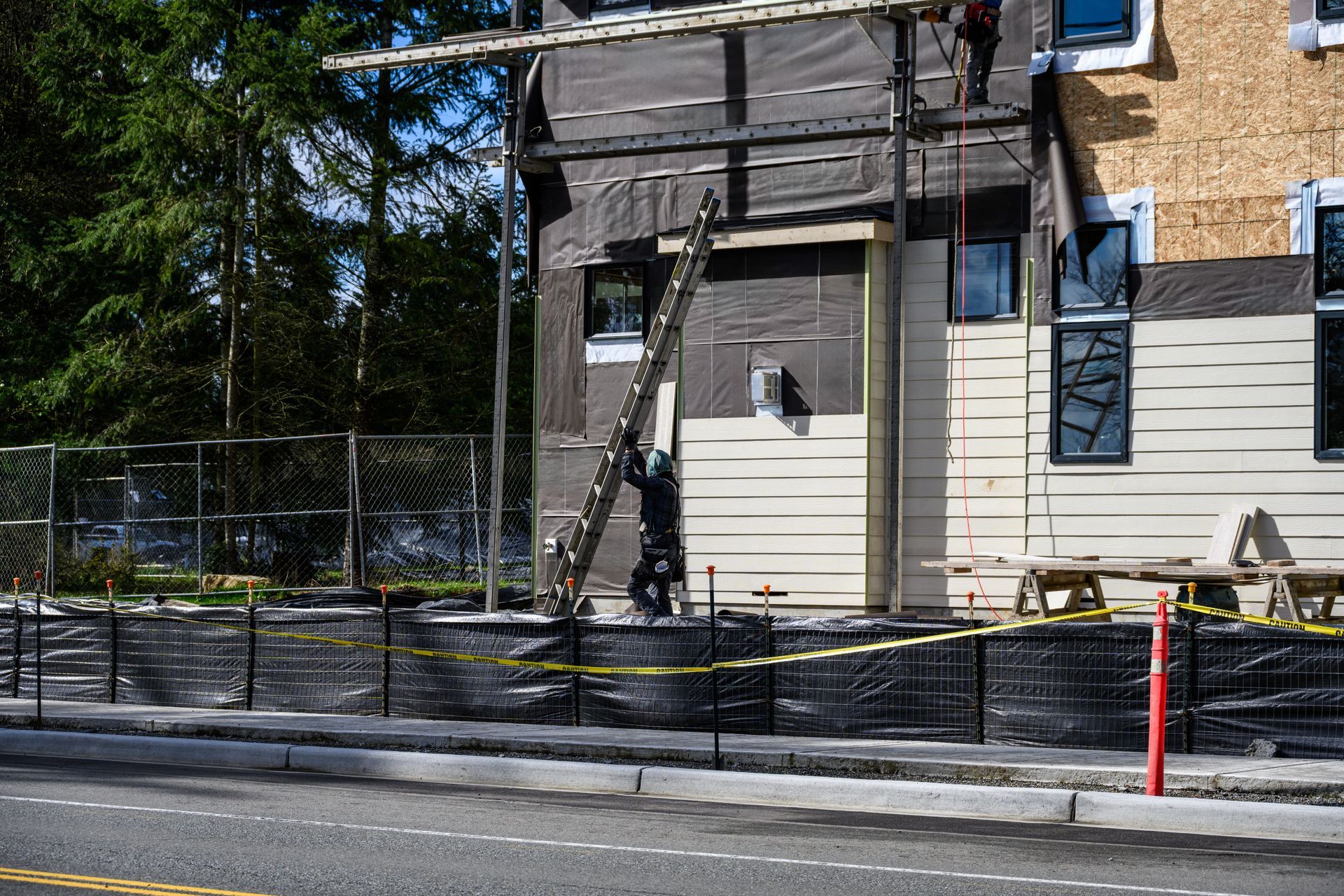 Ouvrier du bâtiment sur une échelle, en train de poser le bardage d'un bâtiment, avec du ruban de signalisation et une route au premier plan.