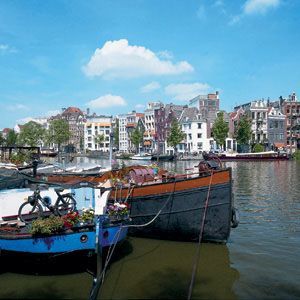 Boats moored on a canal in Amsterdam, with buildings in the background under a blue sky.