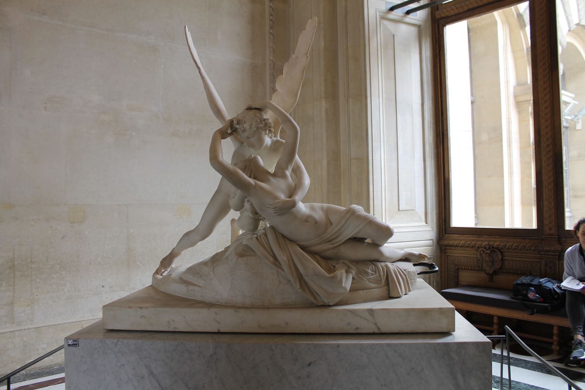 Marble sculpture of a winged figure embracing a reclining figure. Set in a room with a window.