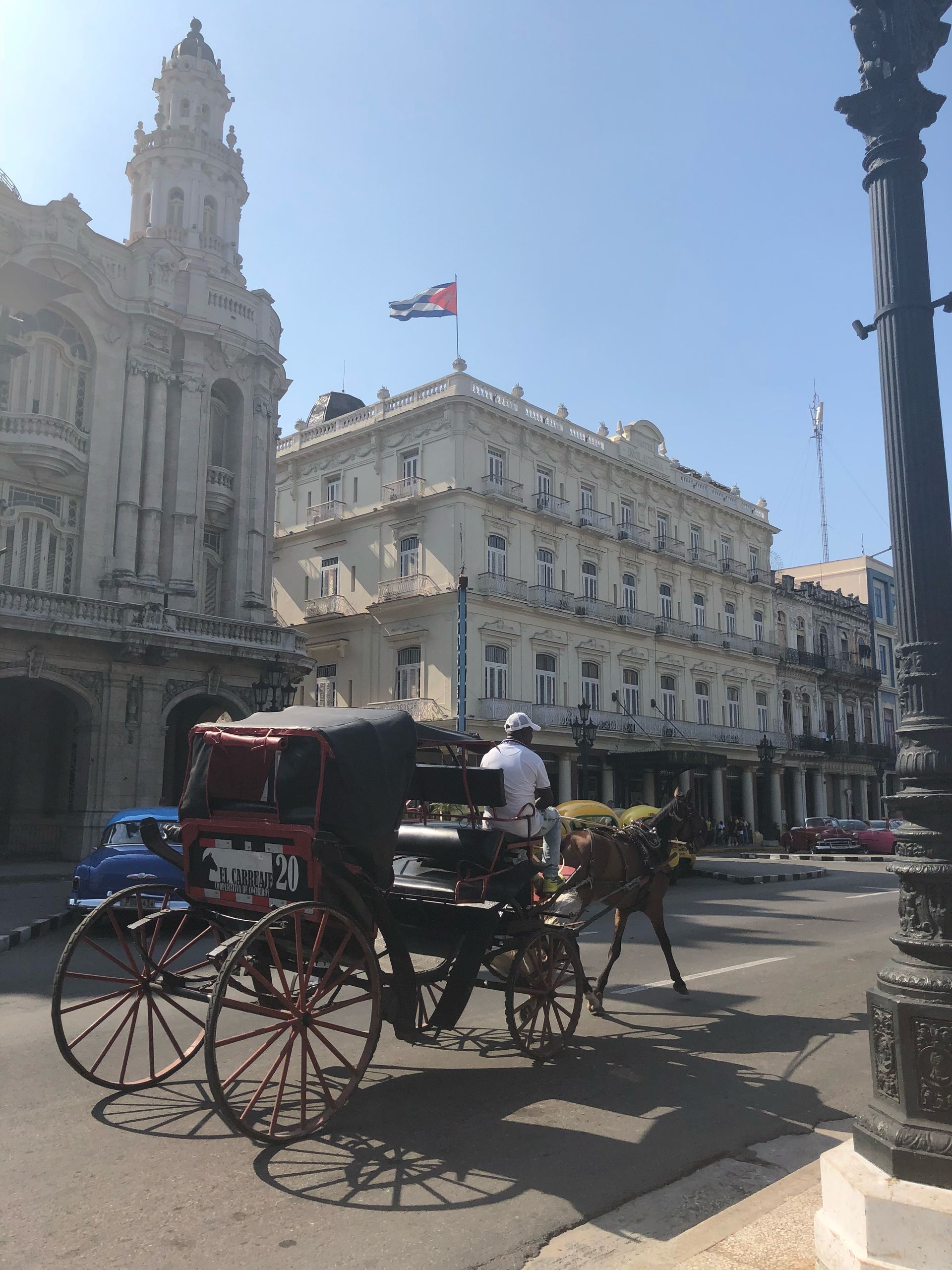 Horse-drawn carriage on a Havana street with ornate buildings, Cuban flag, and blue sky.