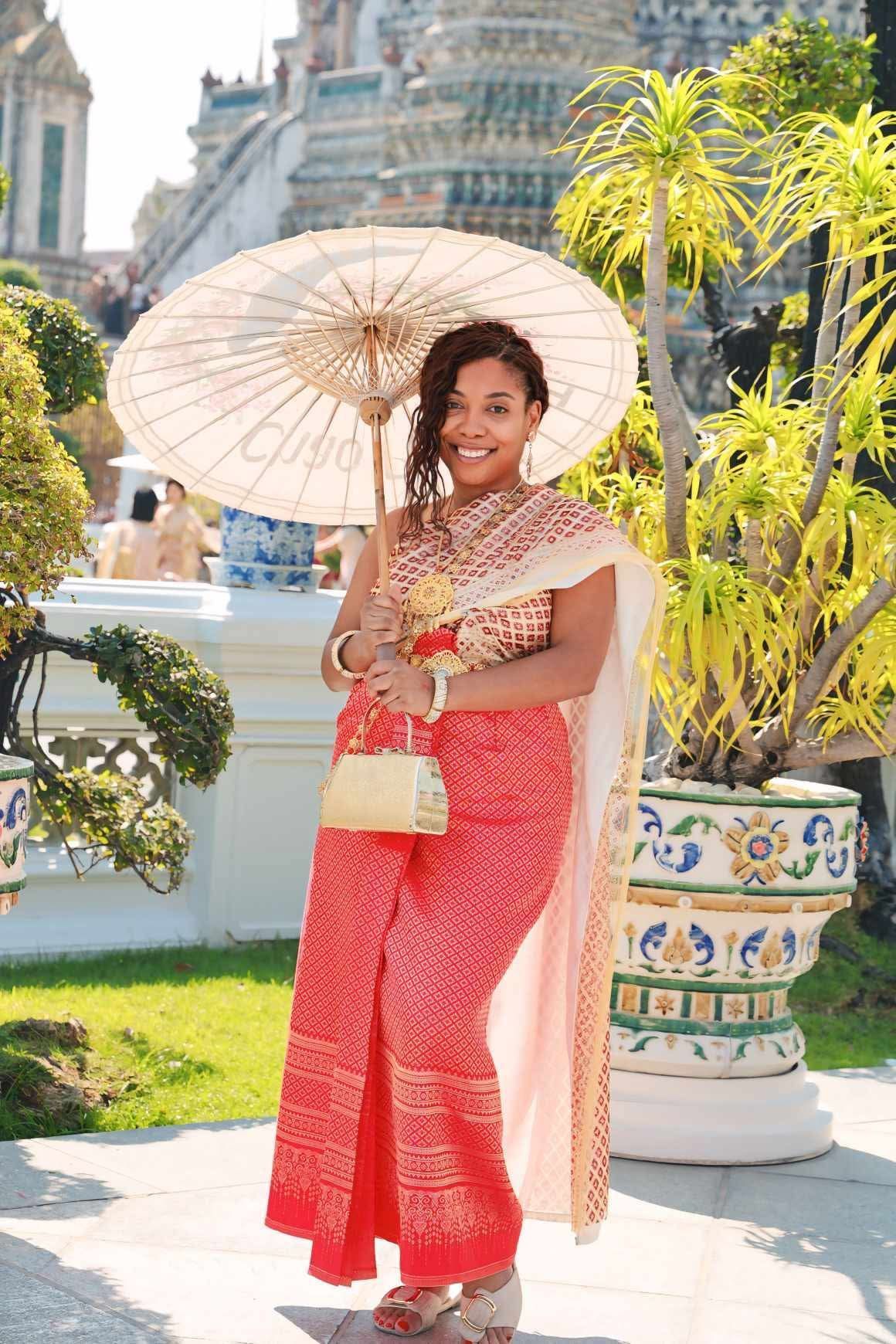 Woman in traditional Thai dress smiles, holds parasol, stands by a temple.