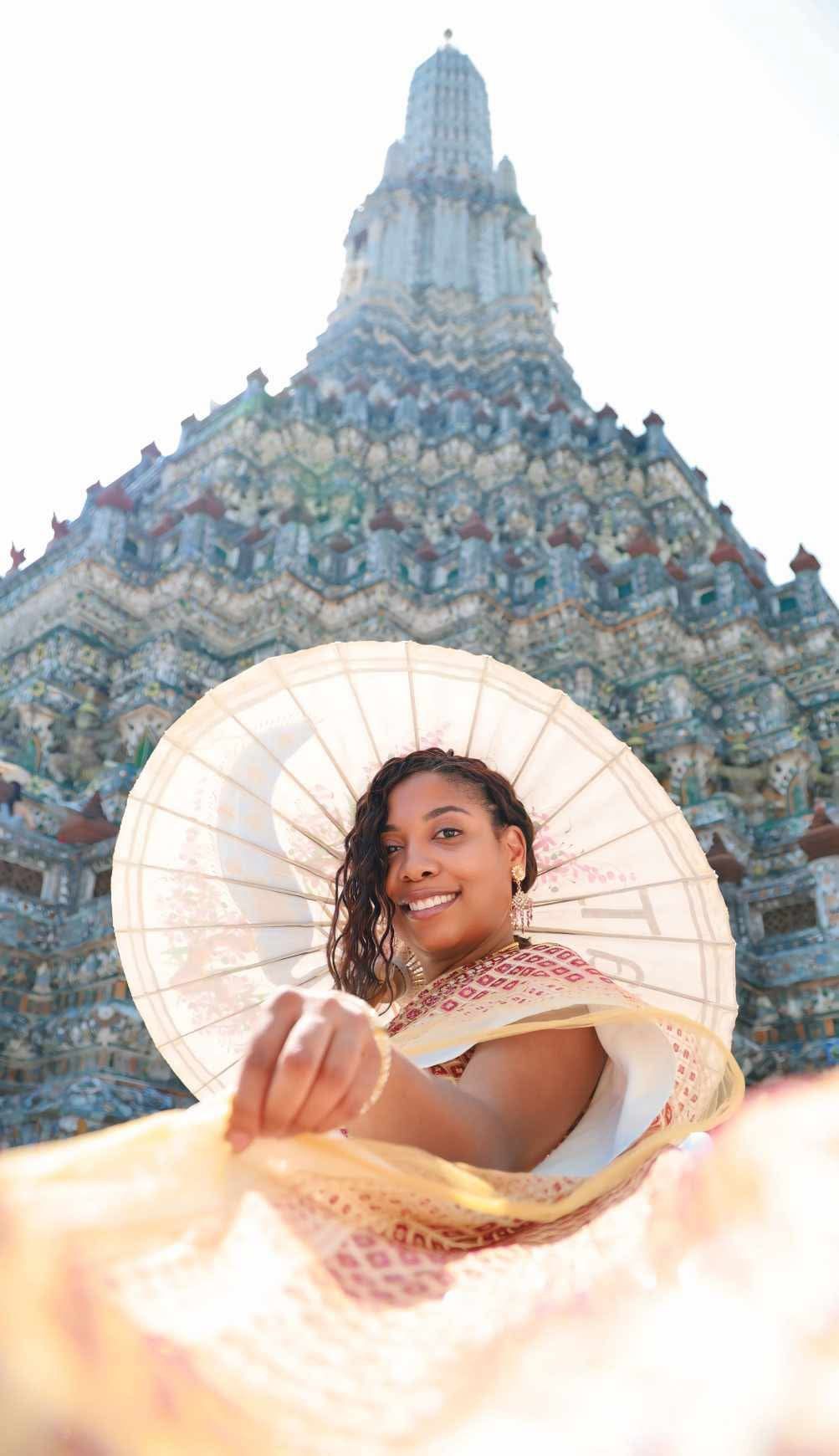 Woman holds parasol in front of Wat Arun temple, smiling, sunny sky.