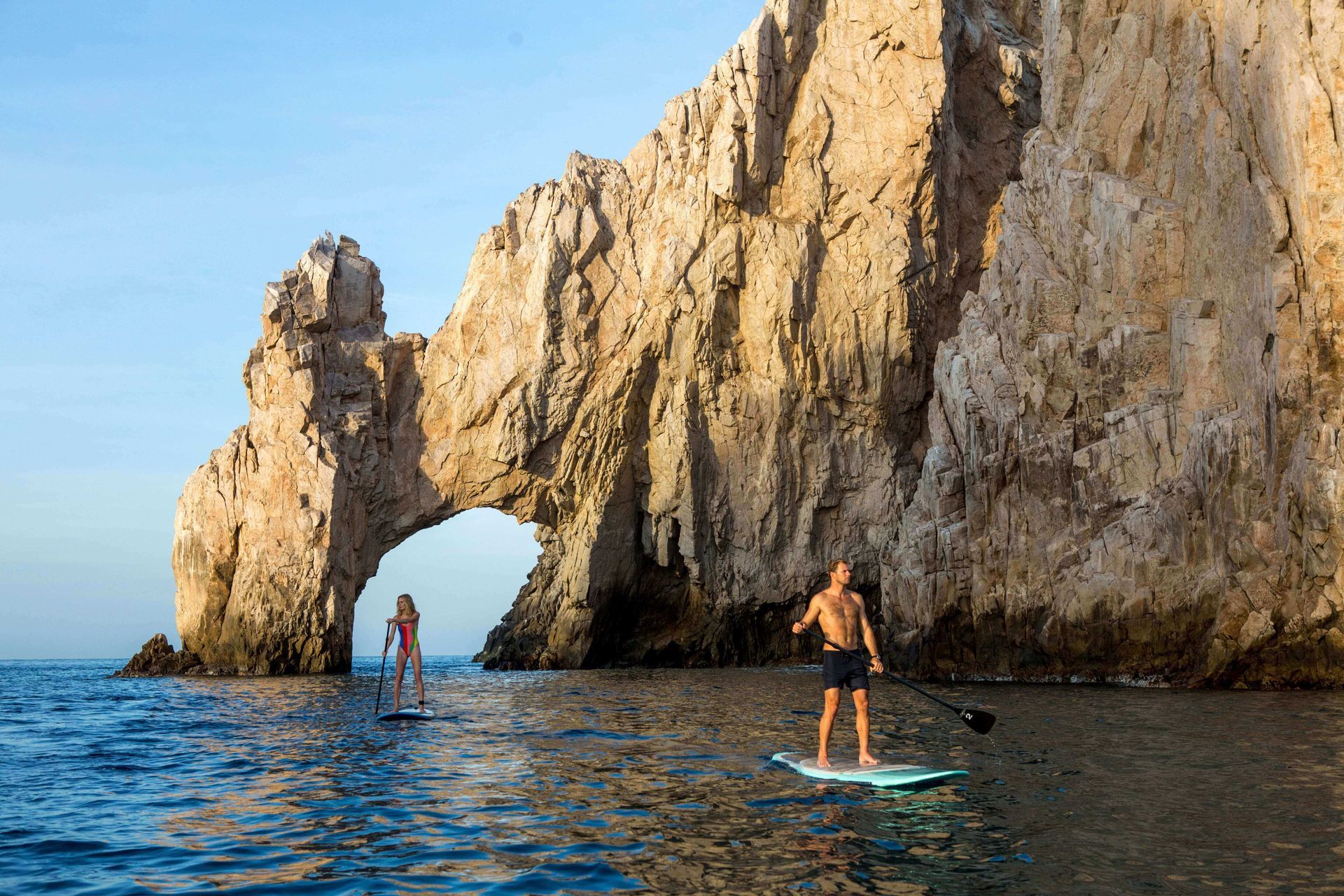 Two people paddleboard near a large rock arch in ocean; sunny, clear sky.