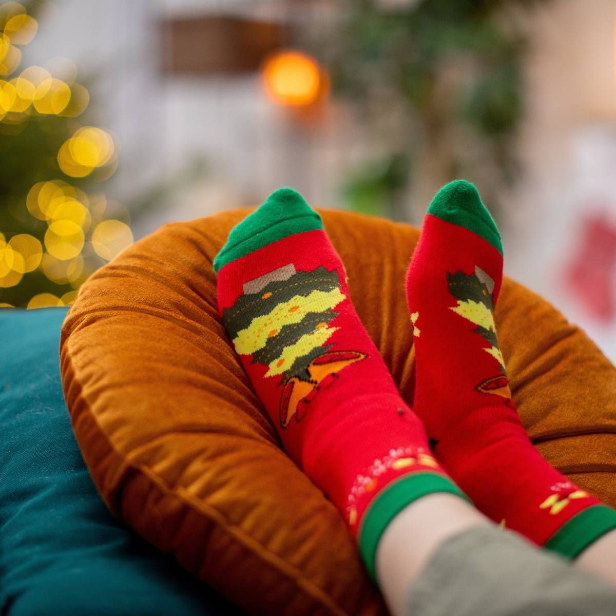 Feet wearing red Christmas socks on an orange cushion, blurred background with lights.