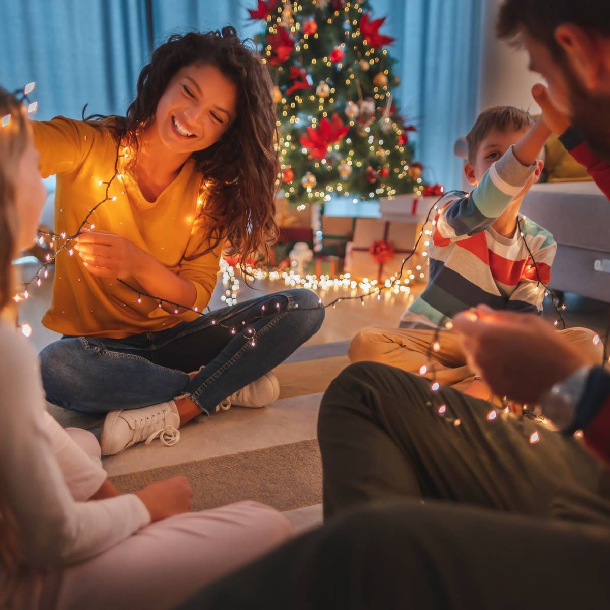 Family laughs while playing with Christmas lights in front of a decorated tree.