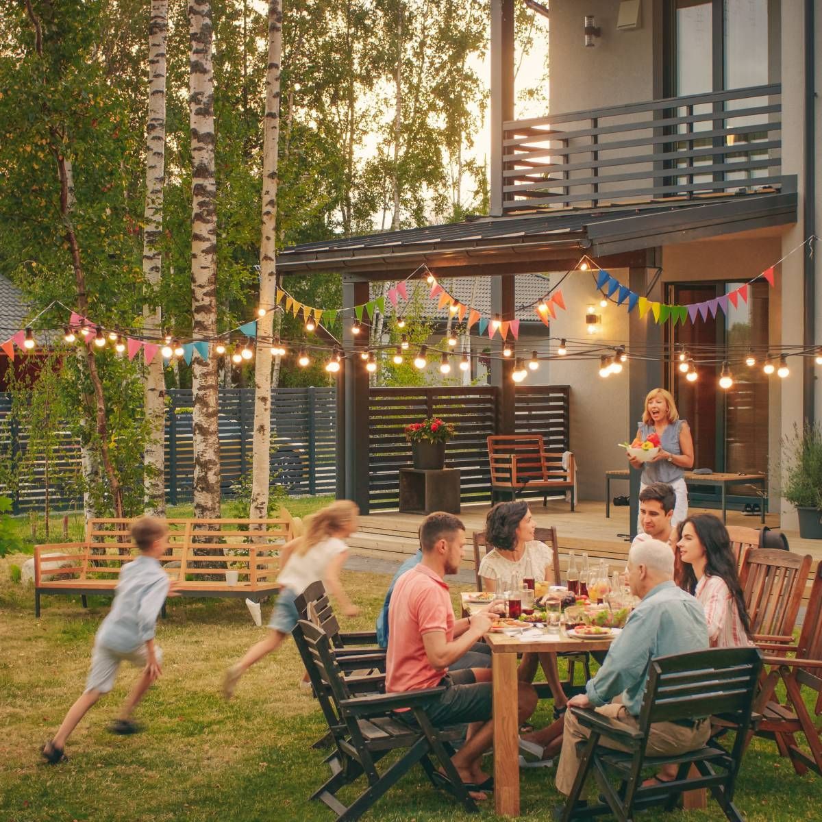Family enjoying a backyard party with food, string lights, and children running.