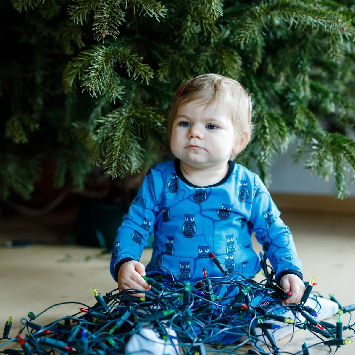 A baby sitting under a Christmas tree, surrounded by tangled string lights, with a curious expression.