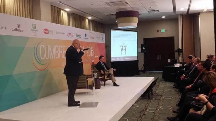 Un orador se dirige a una audiencia desde un escenario en una conferencia, con una pantalla de proyección al fondo.