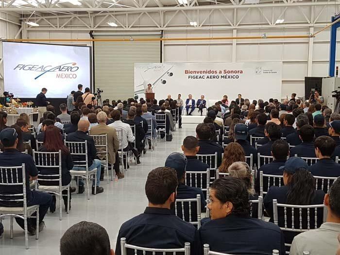 Un numeroso público se sienta en filas de sillas en un evento de Figeac Aero México dentro de una espaciosa nave industrial.