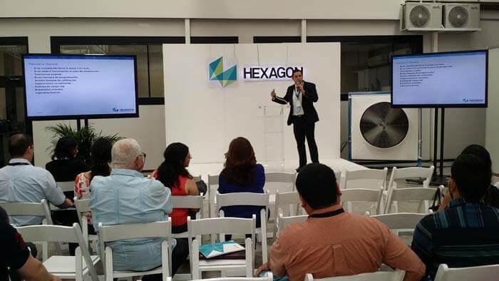 Un orador realiza una presentación en un evento corporativo con el logotipo de la empresa Hexagon detrás y dos pantallas que muestran información.