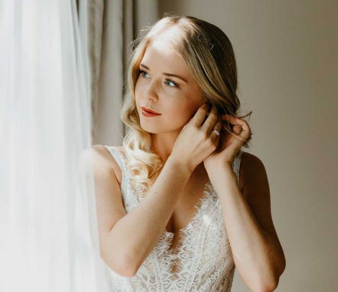 A woman in a wedding dress is putting on her earrings while looking out of a window.