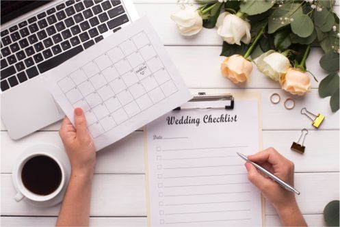 A person is writing on a wedding checklist next to a laptop and a cup of coffee.