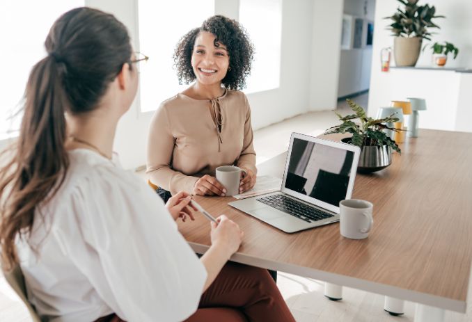 Two women are sitting at a table with a laptop and drinking coffee.