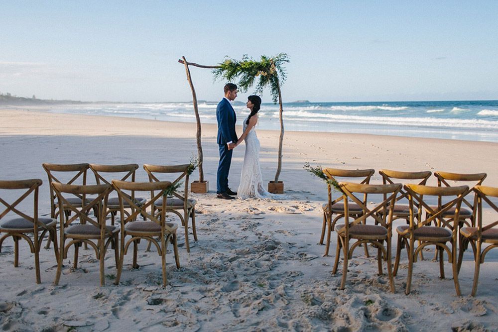 A bride and groom are standing on a beach holding hands.