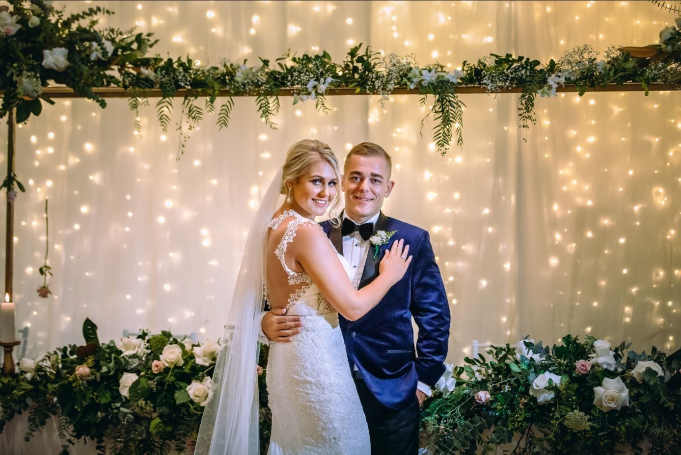 A bride and groom are posing for a picture in front of a curtain of lights.