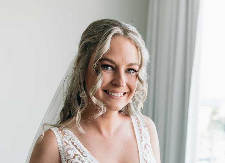 A woman in a wedding dress and veil is smiling in front of a window.