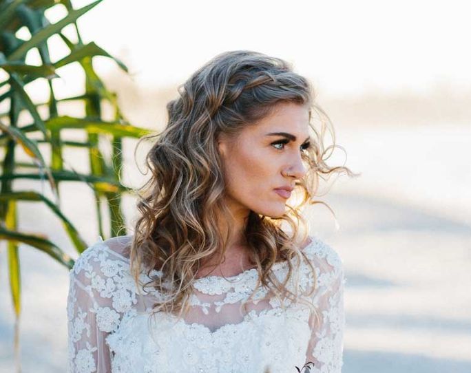 A woman in a wedding dress is standing on a beach next to a palm tree.