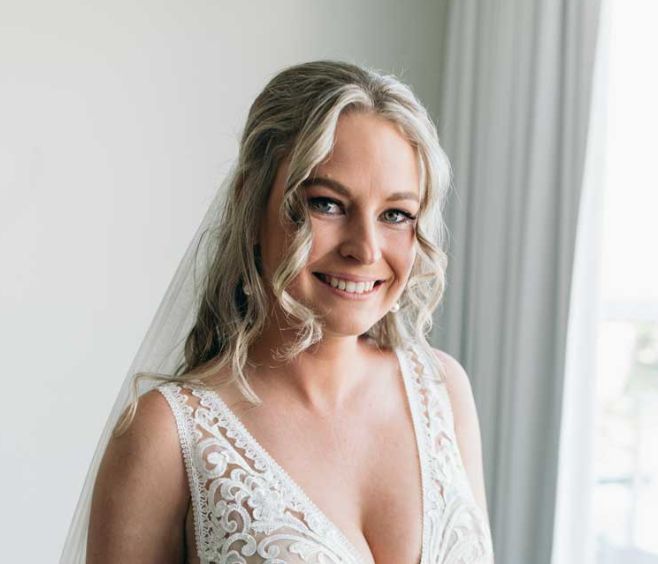 A woman in a wedding dress and veil is smiling in front of a window.