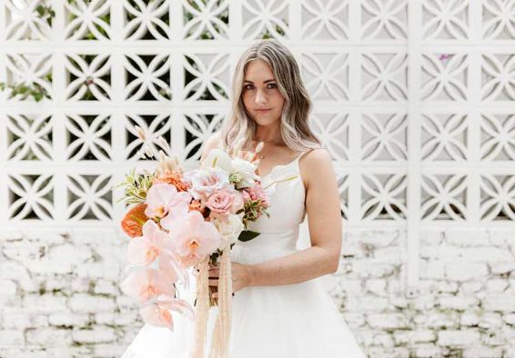 A bride in a white dress is holding a bouquet of flowers in front of a white wall.