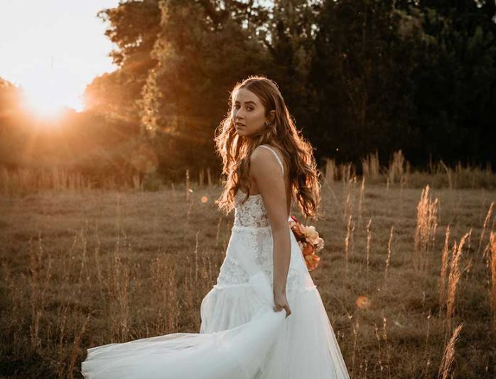 A woman in a wedding dress is walking through a field at sunset.