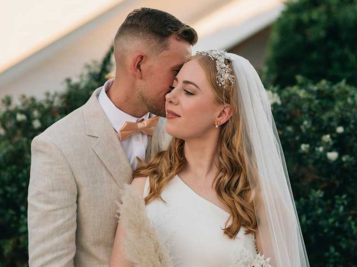 A bride and groom are posing for a picture and the groom is kissing the bride on the forehead.