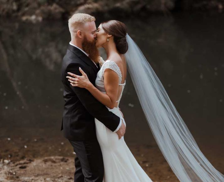 A bride and groom are kissing in front of a body of water.