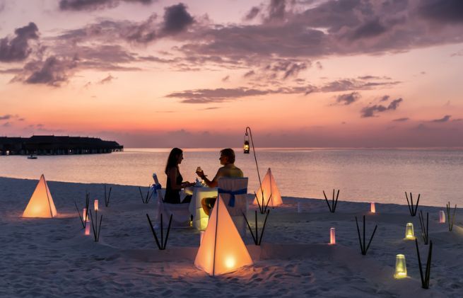 A man and a woman are sitting at a table on the beach at sunset.