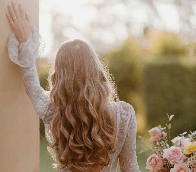 A woman in a white dress is standing next to a wall holding a bouquet of flowers.