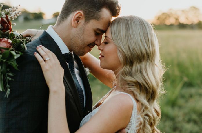 A bride and groom are touching their foreheads in a field.