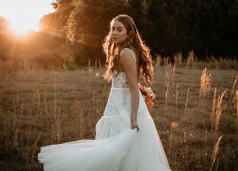 A woman in a wedding dress is standing in a field at sunset.