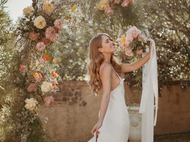 A woman in a white dress is standing in front of a floral arch.