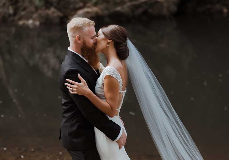 A bride and groom are kissing in front of a body of water.