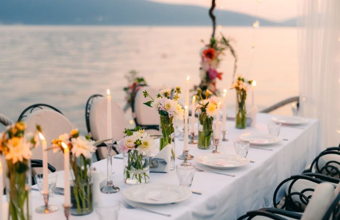 A long table with plates , candles , vases of flowers and a view of the ocean.