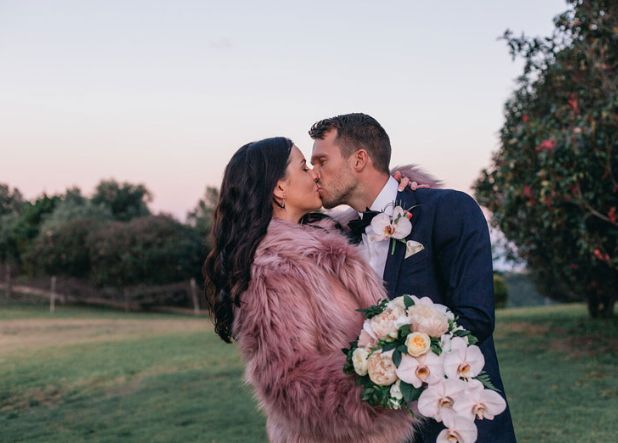 A bride and groom are kissing in a field . the bride is wearing a pink fur coat.