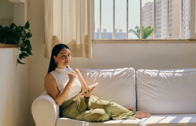 A woman is sitting on a couch holding a book and a pen.