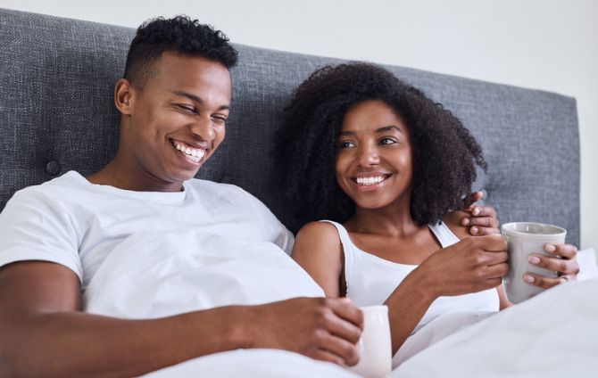 A man and a woman are laying in bed holding cups of coffee.