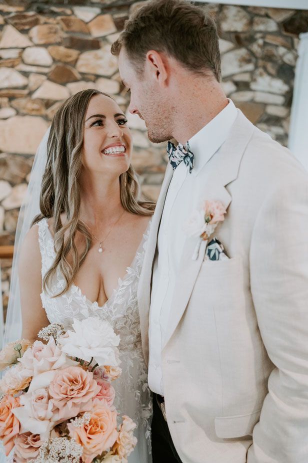 A bride and groom are looking at each other in front of a stone wall.