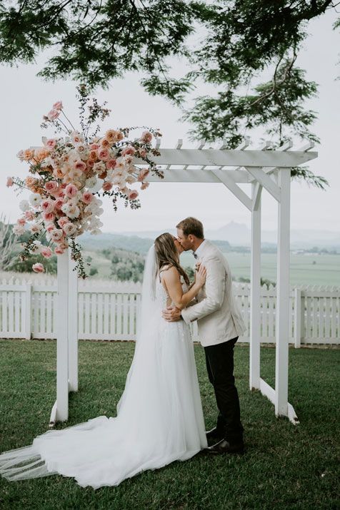 A bride and groom are kissing under a white arch at their wedding.