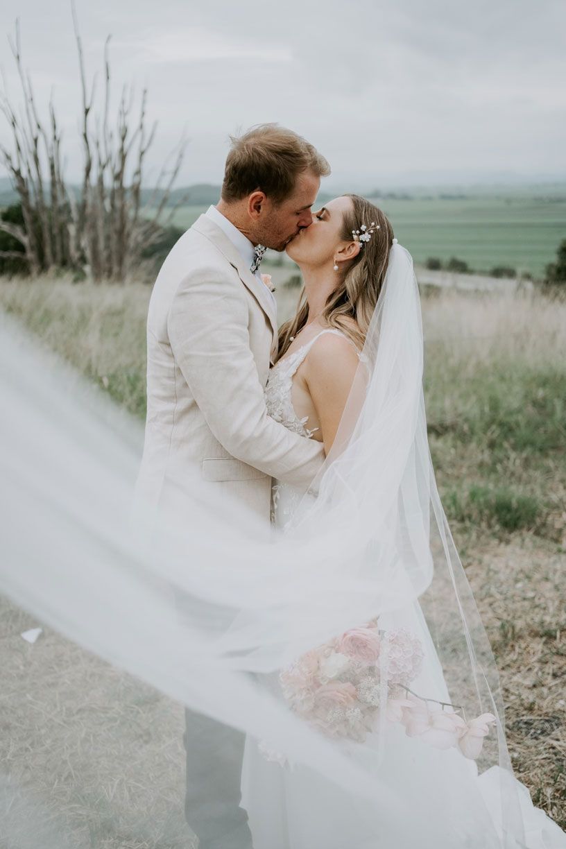 A bride and groom are kissing under a veil that is blowing in the wind.