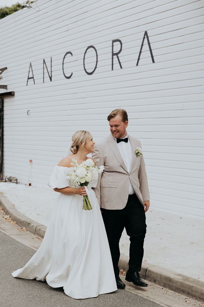 A bride and groom are walking down the street in front of a white building.