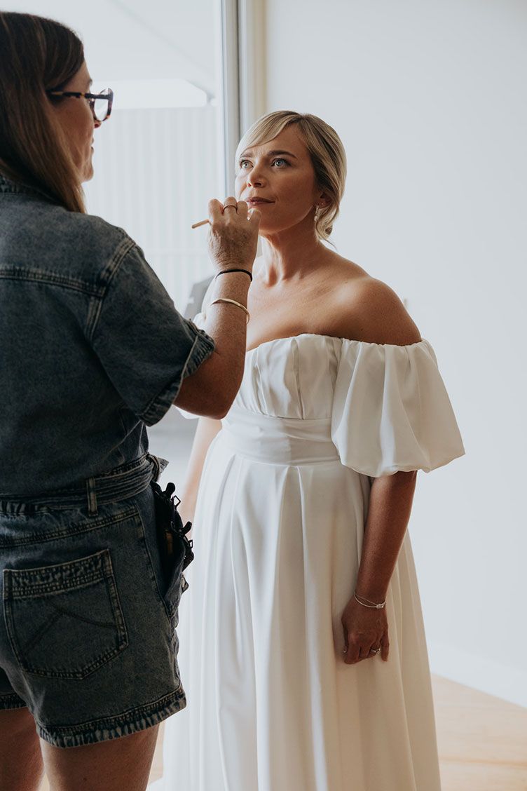 A woman in a white dress is getting her makeup done by a makeup artist.