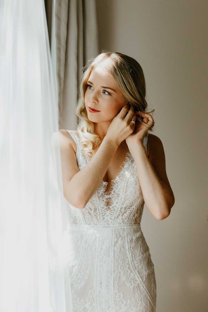 A woman in a wedding dress is putting on her earrings while standing in front of a window.