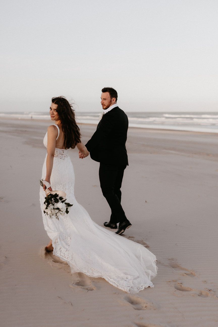 A bride and groom are walking on the beach holding hands.