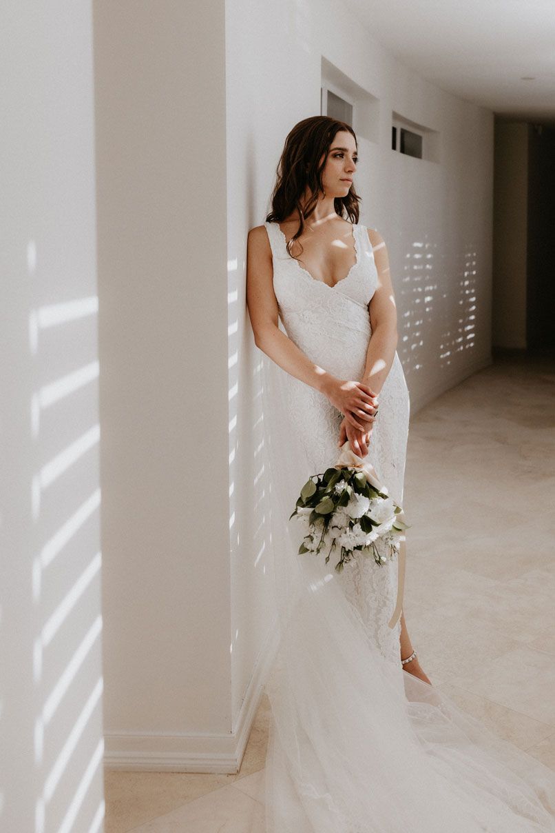 A woman in a white dress is leaning against a wall holding a bouquet of flowers.