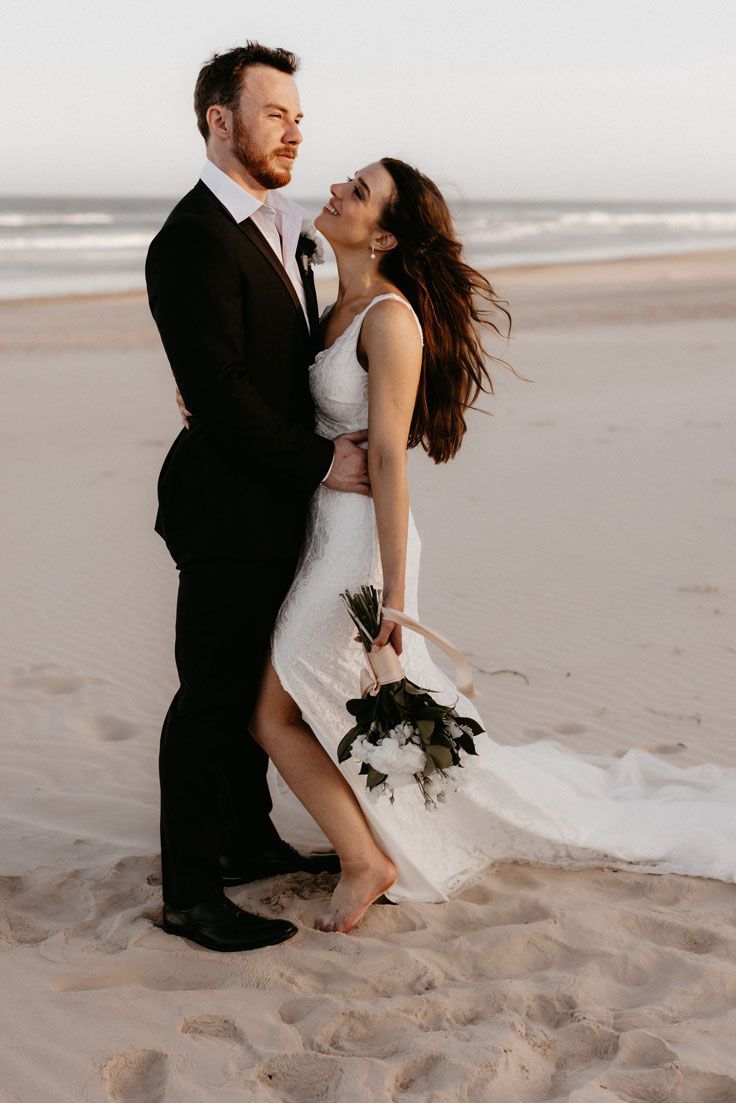 A bride and groom are posing for a picture on the beach.