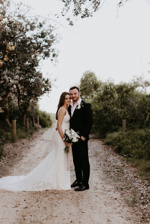 A bride and groom are standing next to each other on a dirt road.