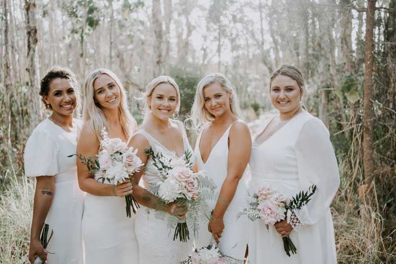 The bride and her bridesmaids are posing for a picture in the woods.