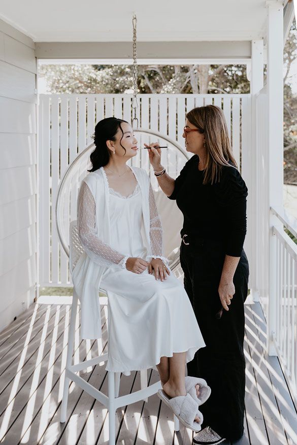 A woman is applying makeup to a woman sitting in a chair on a porch.