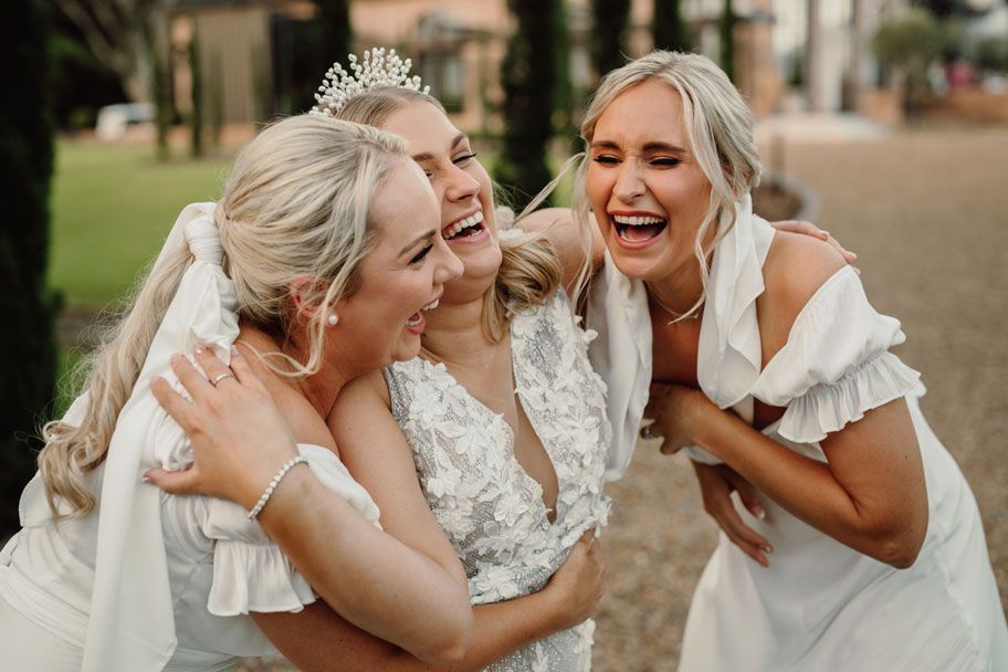 Three women in white dresses are hugging each other and laughing.