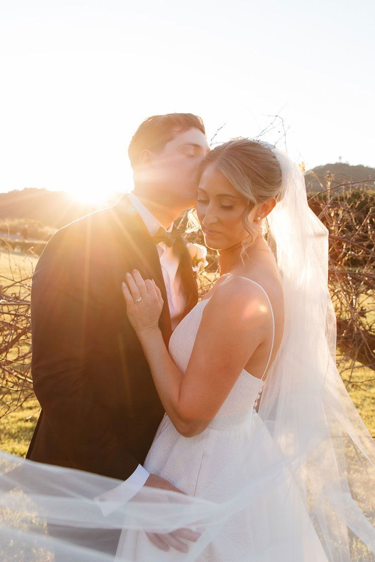 A bride and groom are kissing in a field with the sun shining through the veil.